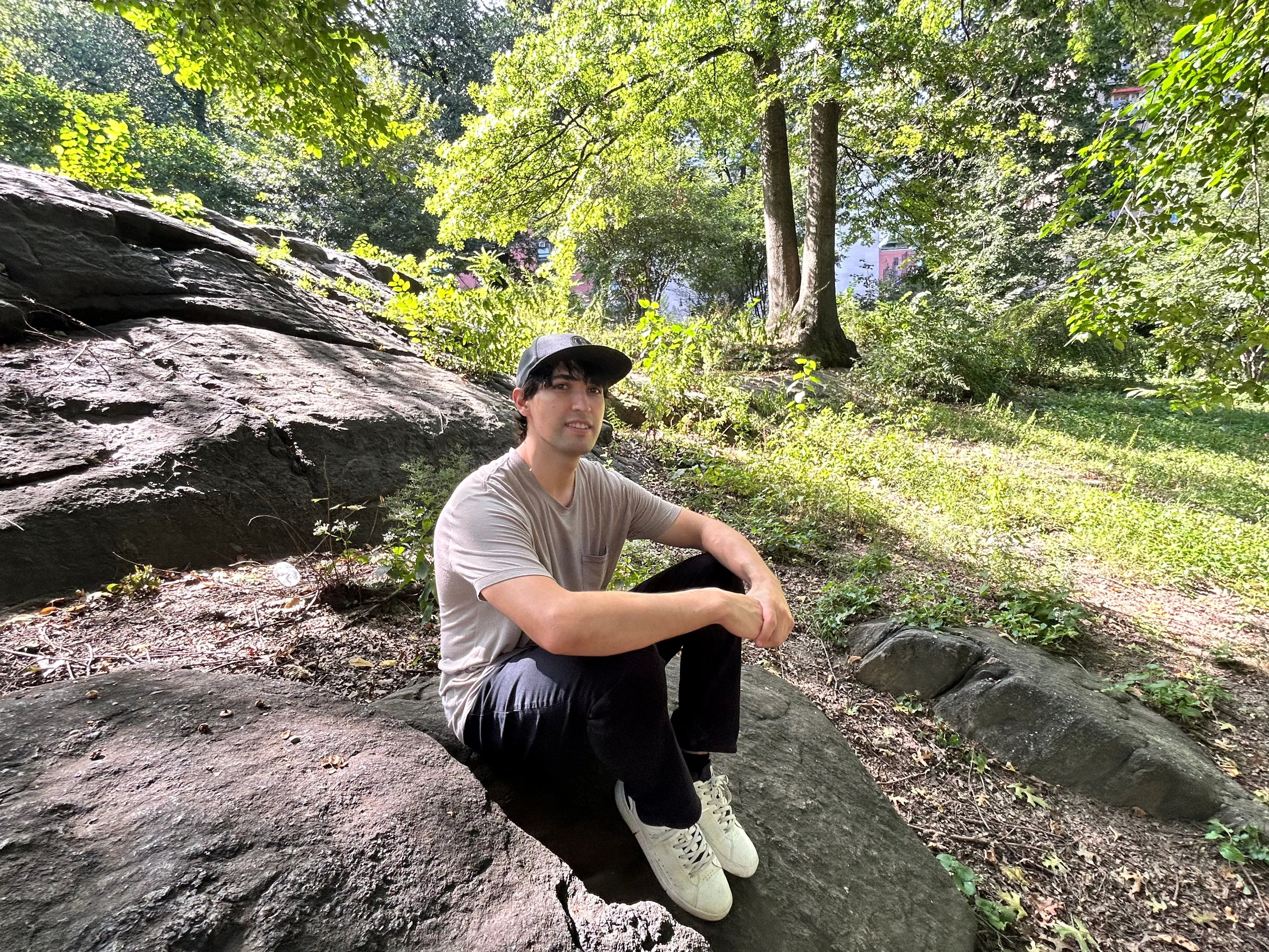 Jordan sitting on a rock outcrop in a city park, in a cap and a pale t-shirt, with sunlit trees and grass behind him.
