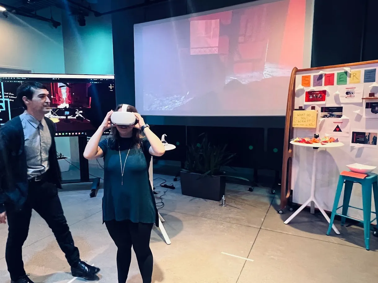 A showcase floor. Someone stands centre with both hands on a white VR headset, the Soteria scene projected on the wall behind them. Jordan walks past on the left. To the right, a hand-made display board spells out SOTERIA in cut-out letters above pinned artifacts, a sticky note asking what do you wish, and a bowl of folded paper boats.