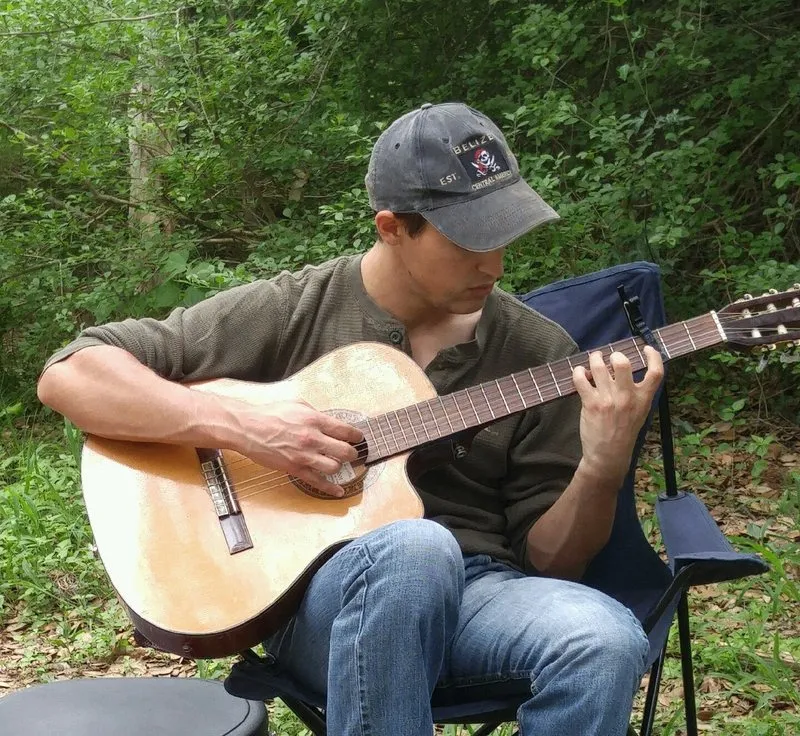 Jordan playing a classical guitar, seated outdoors in a camp chair with woodland behind him.